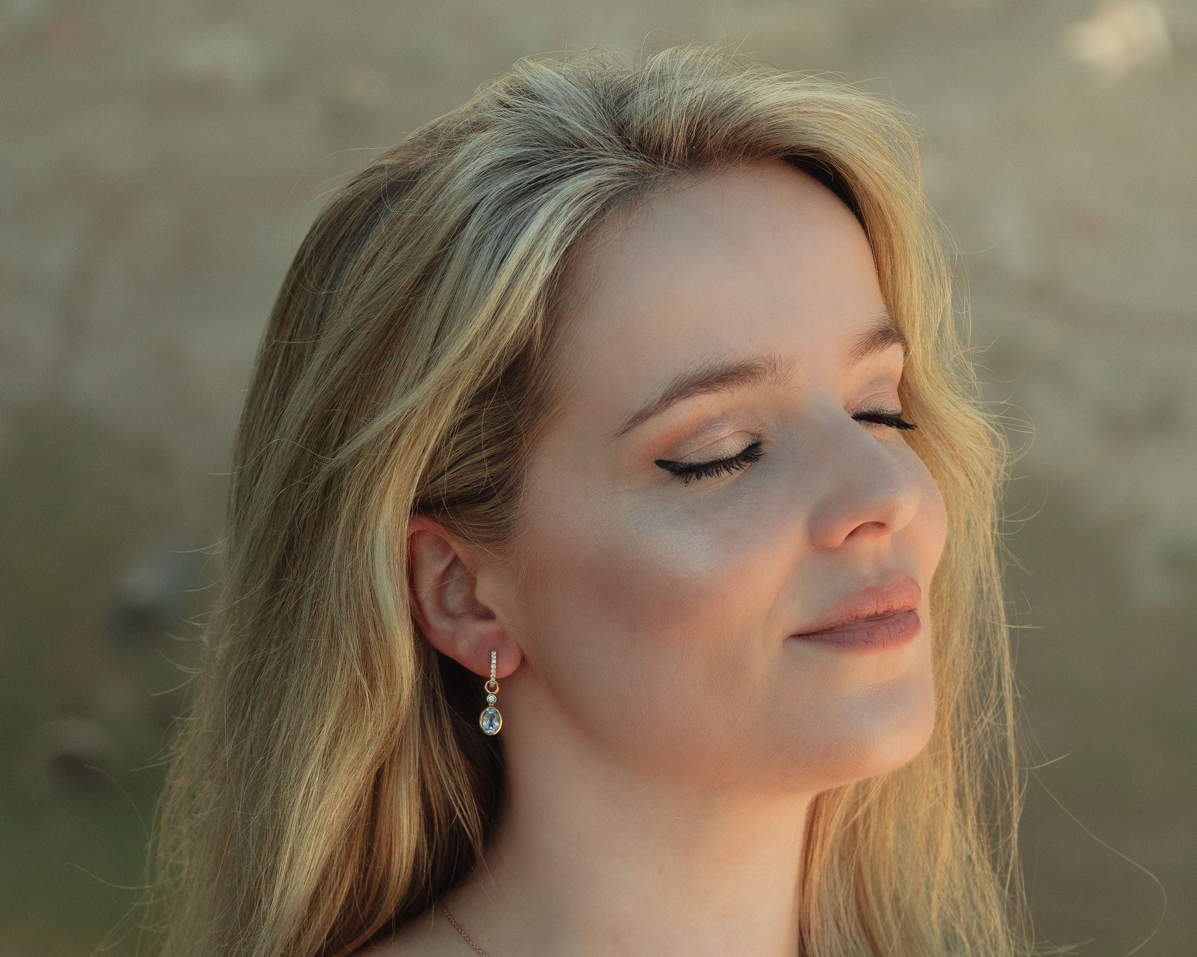 Woman with long blonde hair wearing a LUNA 18ct yellow gold and diamond huggie earrings with blue topaz and diamond earring attachments with a blurred natural background
