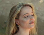 Woman with long blonde hair wearing a LUNA 18ct yellow gold and diamond huggie earrings with blue topaz and diamond earring attachments with a blurred natural background